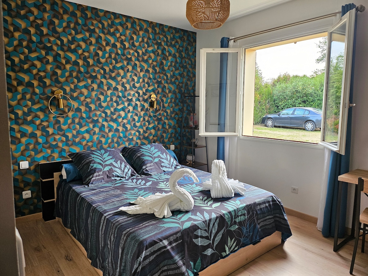 A stylish bedroom features a patterned accent wall in shades of blue and gold. A spacious bed is adorned with a dark floral duvet. Two swan-shaped towels are artfully arranged on the bed, and a window offers natural light and a view of the greenery outside.
