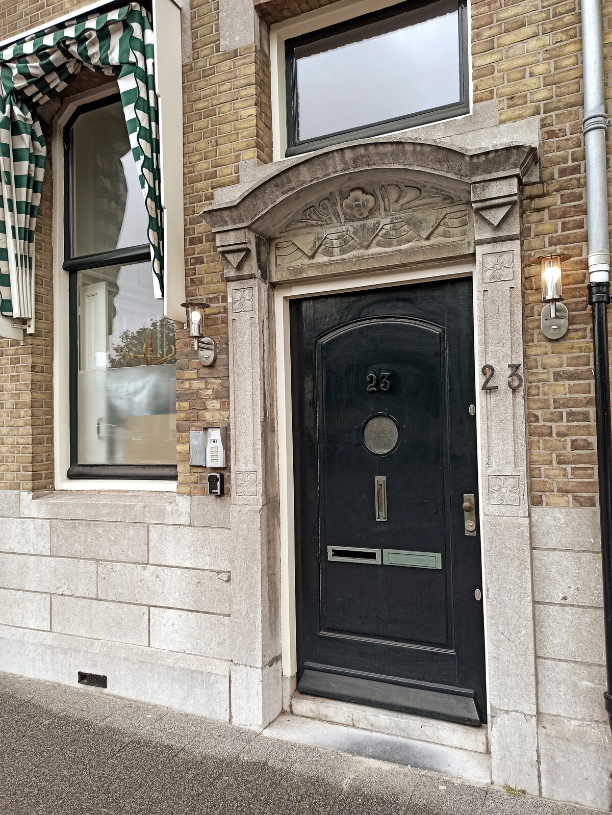 The entrance to a historic building is showcased, featuring a dark wooden door with an oval glass panel and ornate detailing above. The exterior is complemented by light brickwork and green-and-white striped awnings over large windows on either side.