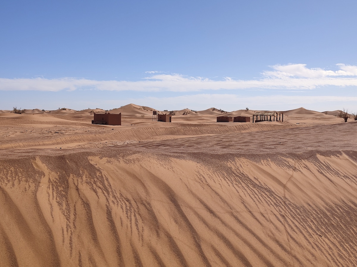 The image captures a serene desert landscape with gentle sand dunes, showcasing the rich textures of the golden sand. In the distance, a few simple structures are visible, blending harmoniously with the natural surroundings under a clear blue sky.