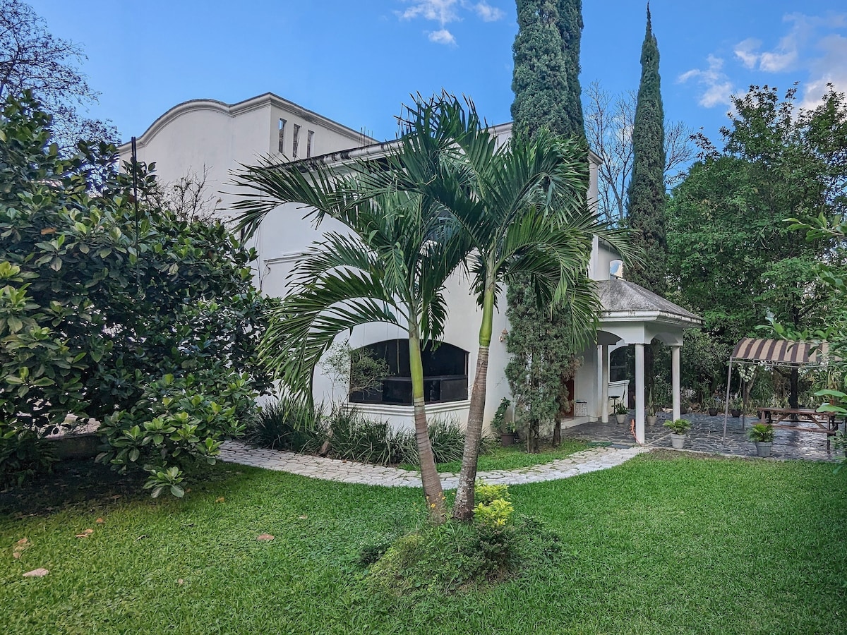 A sprawling garden features well-maintained grass and tropical plants. A single palm tree adds height to the landscape. The exterior of a multi-story house is visible, with large windows providing views of the surroundings. A stone pathway leads to a shaded seating area.