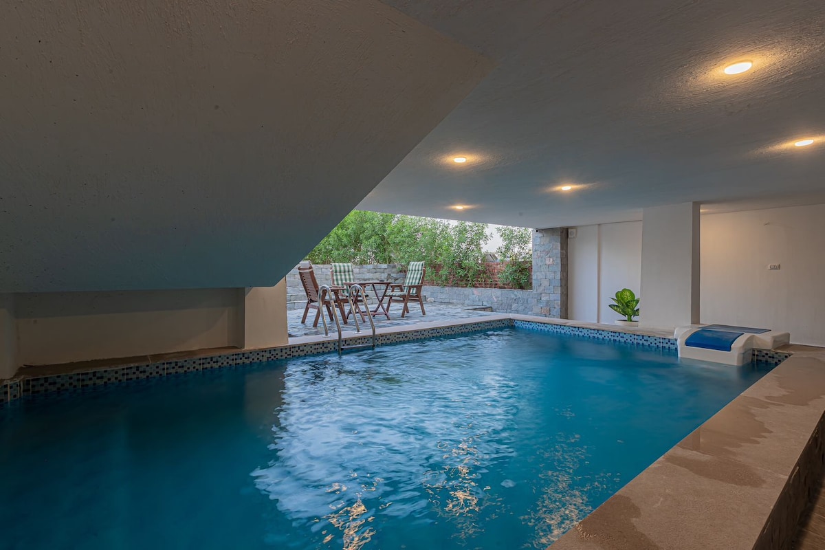 An indoor pool glistens under soft ceiling lights, surrounded by neutral-toned walls. The space features a set of wooden chairs placed on a deck outside, providing a view of the greenery beyond. A lounge area is visible, enhancing relaxation options.