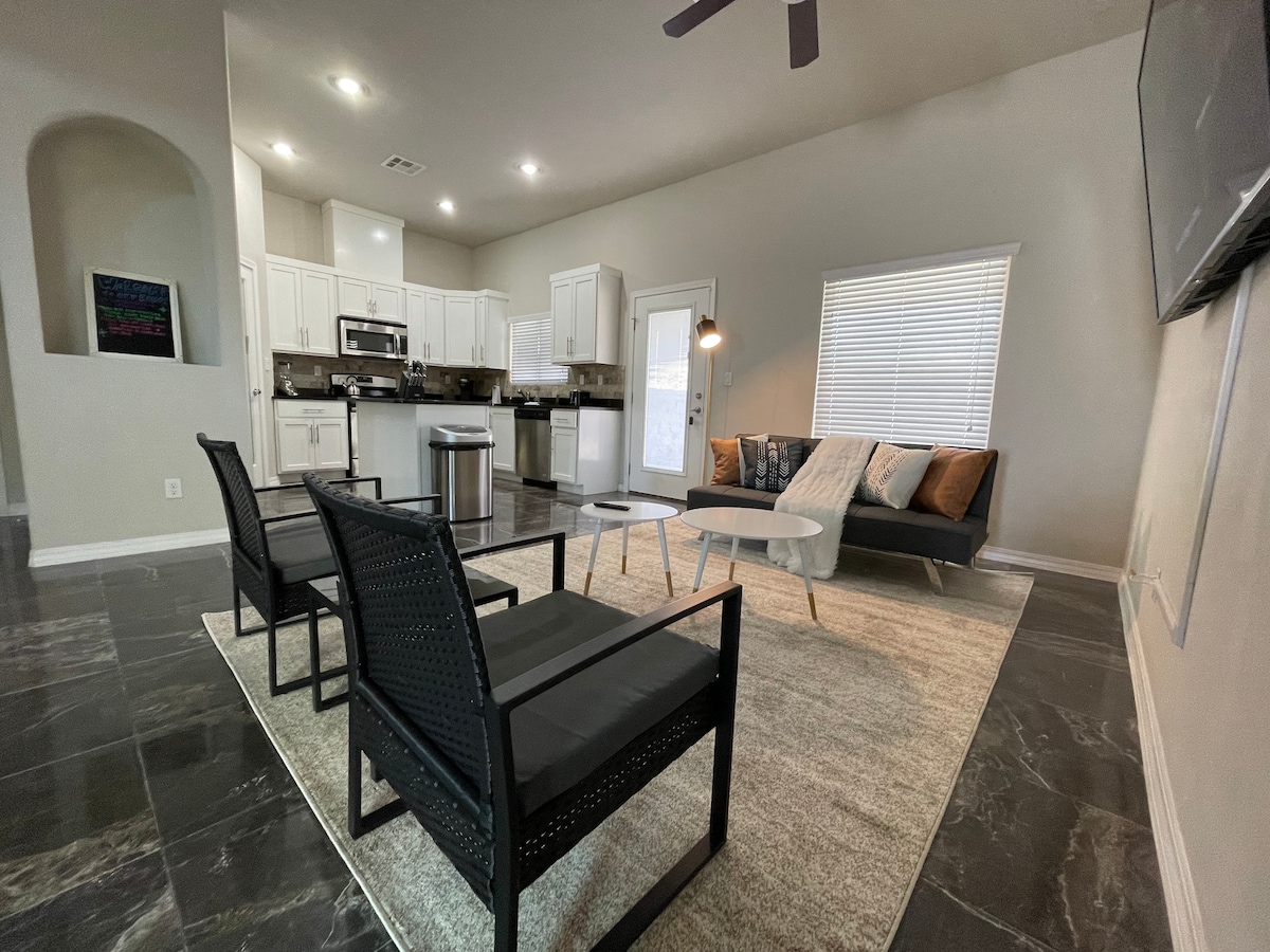 The open living area features modern furnishings, including a sleek black sofa and minimalist coffee table, arranged on a neutral-toned area rug. Natural light enters through a window, illuminating the dining area and kitchen in the background, creating a bright and airy atmosphere.