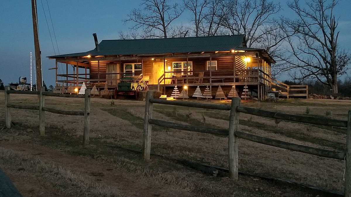 The rustic cabin is illuminated in the evening, showcasing its wooden exterior and wrap-around porch. The structure is surrounded by open grassland and a split-rail fence, with trees in the background and subtle lighting highlighting its features.
