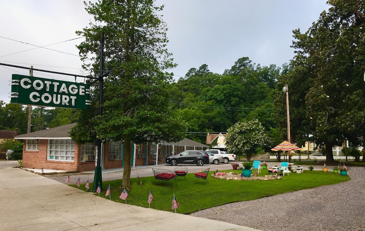 The exterior of Cottage Court is depicted, featuring a vintage sign and vibrant landscaping. A gravel parking area accommodates multiple vehicles, while colorful chairs and umbrellas are arranged around a stone fire pit. Lush trees and greenery frame the scene, creating a welcoming environment.