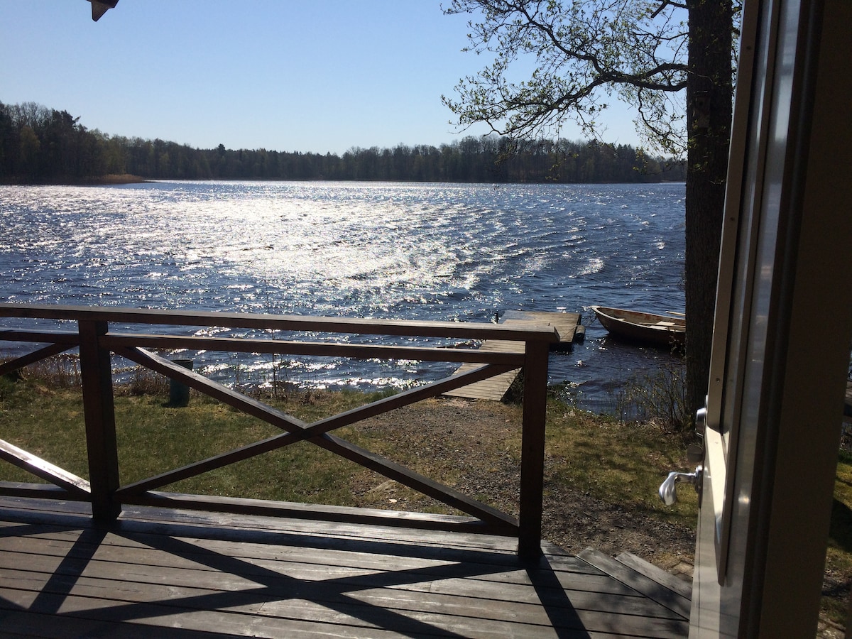 A view of the shimmering lake is presented from a wooden deck, with gentle waves reflecting sunlight. A wooden dock extends into the water, and a small boat rests nearby, surrounded by greenery and trees in the background, enhancing the serene lakeside ambiance.