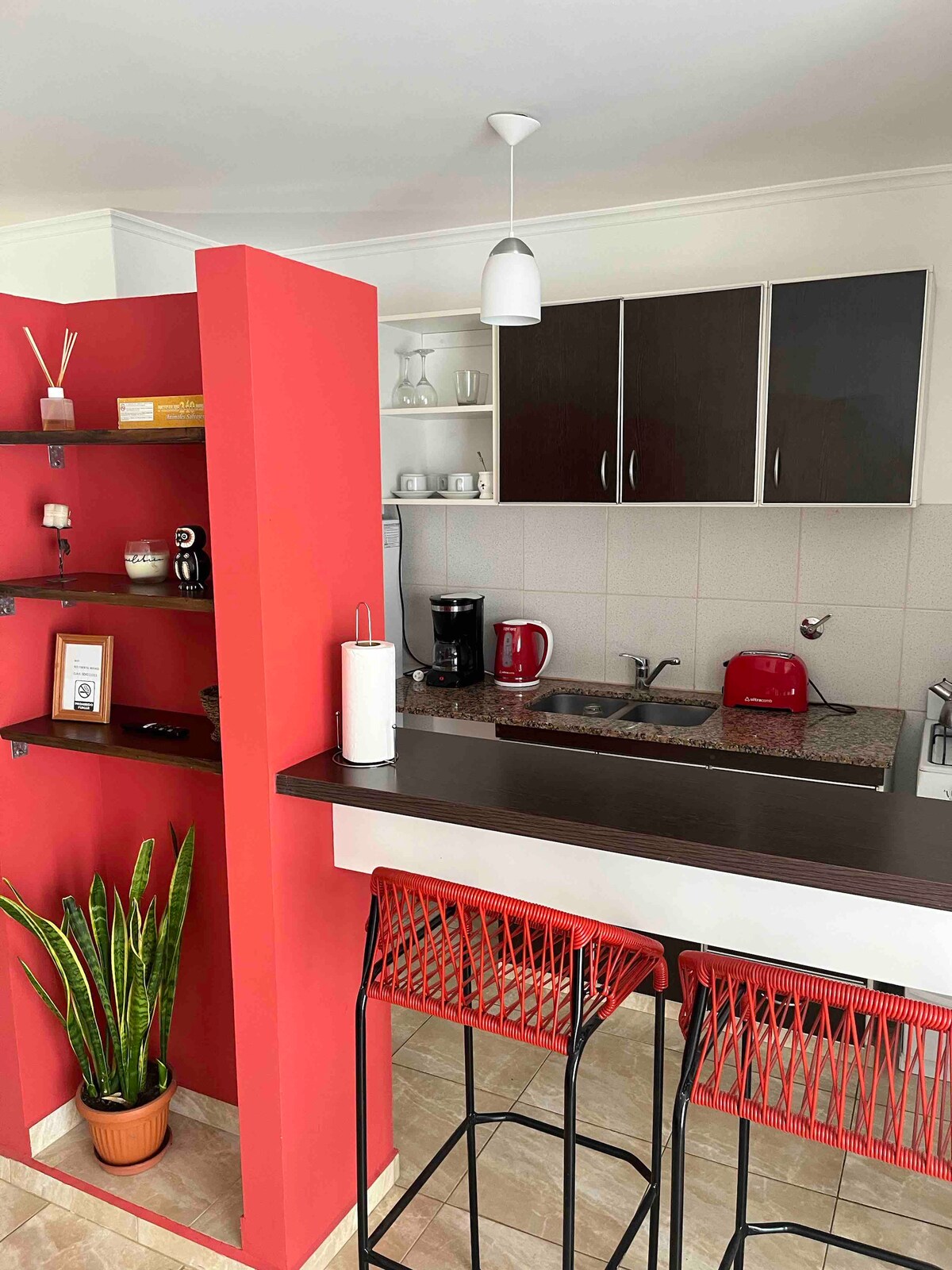 The kitchen area features a red accent wall and a functional layout. Modern cabinetry holds dishware, while kitchen appliances in red and black complement the design. Bar stools in a woven pattern are positioned by the counter, creating a casual dining space. A green plant adds a touch of nature.