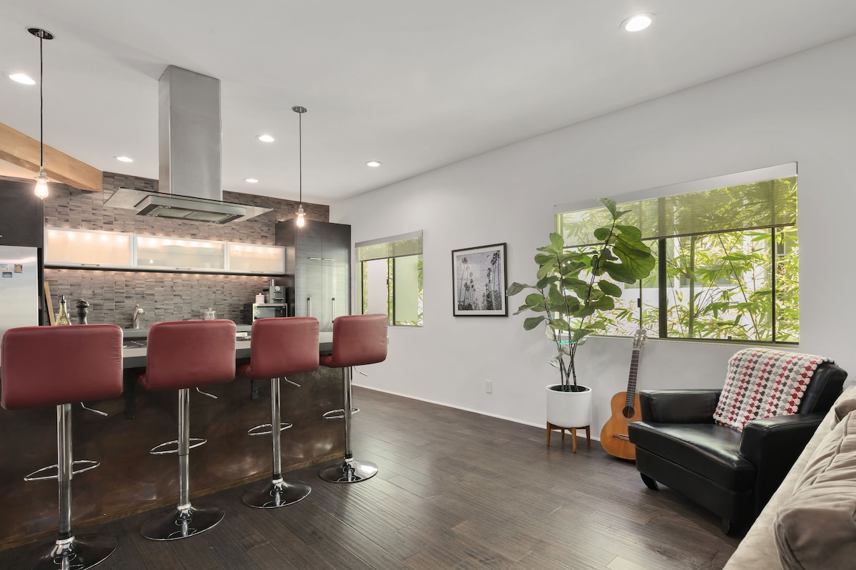 The modern kitchen is equipped with stainless steel appliances and a sleek island with four red barstools. Natural light floods the space through large windows, highlighting a cozy seating area with a black armchair, a decorative plant, and a guitar in the corner.