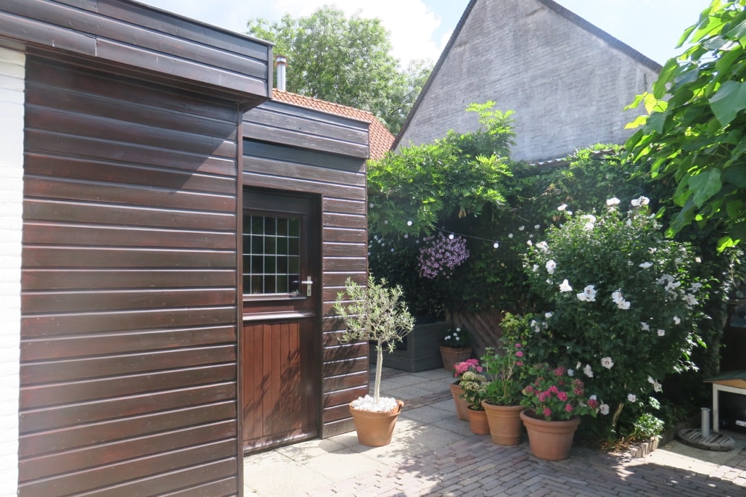 The exterior of the guest suite is shown, featuring wooden paneling and a door with a window. In the courtyard, potted plants with blooming flowers enhance the natural ambiance, with greenery climbing along the walls, creating a serene and inviting outdoor space.
