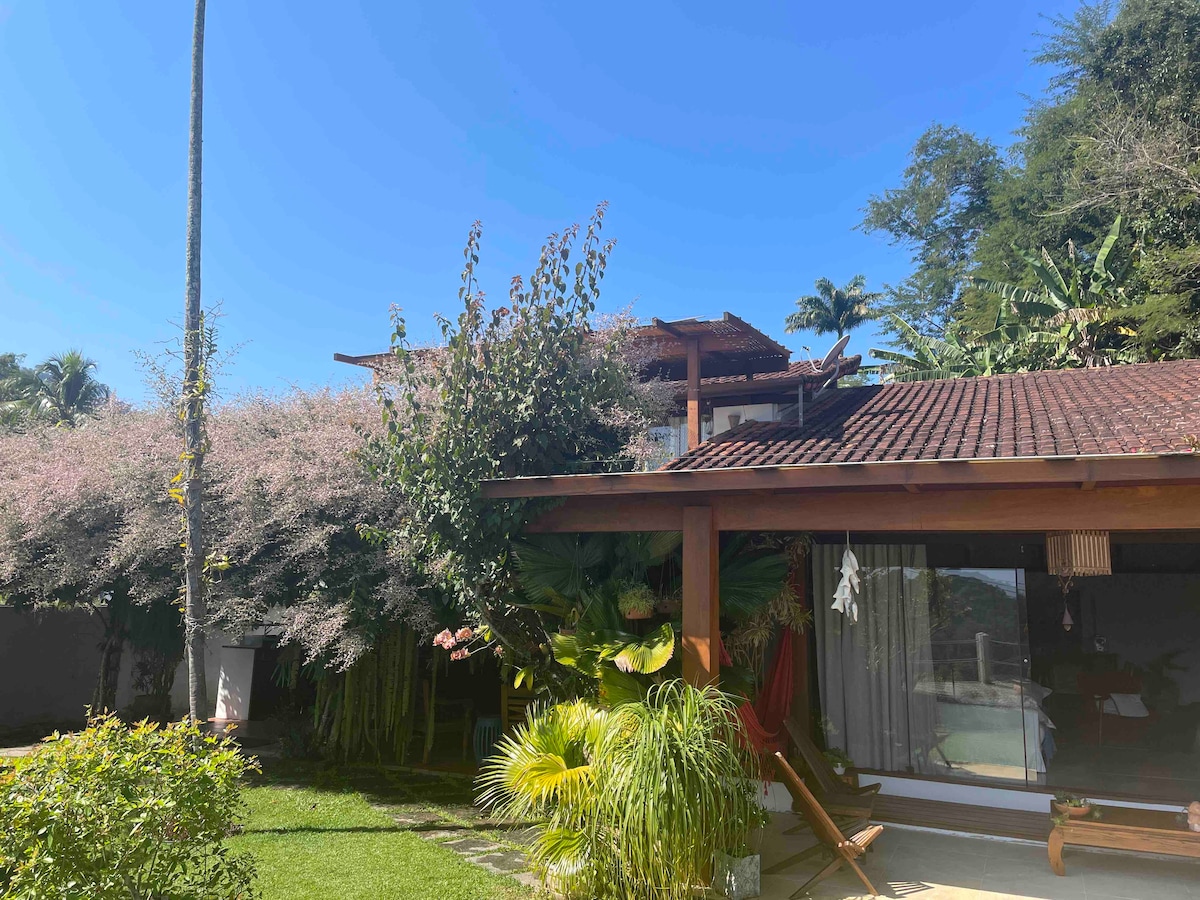 The exterior of the house is surrounded by lush greenery and flowering plants, providing a serene atmosphere. A wooden deck is visible with comfortable lounge furniture, while large windows reflect the inviting interior. The blue sky complements the tranquil setting.