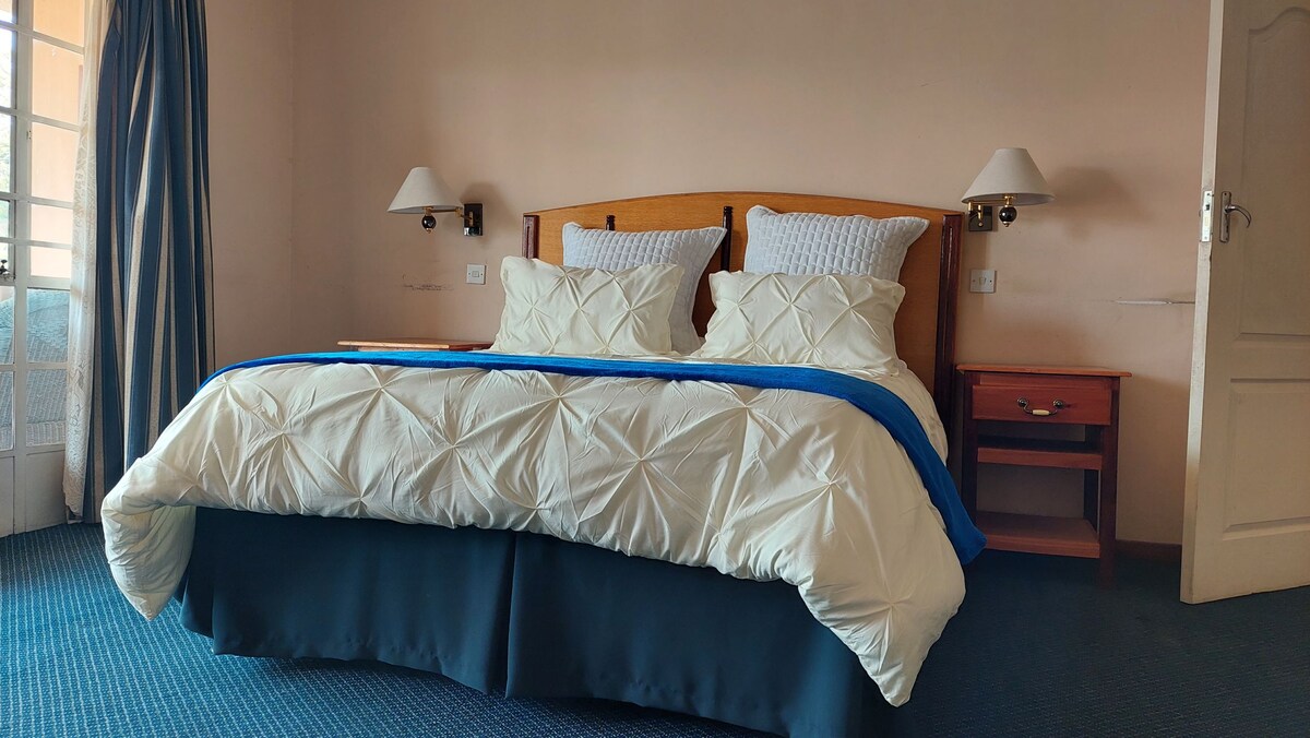 A spacious bed with a tufted white comforter and blue throw is positioned against a wooden headboard. Nightstands are located on either side, equipped with lamps. Natural light enters through a nearby balcony door, enhancing the calm atmosphere of the room.