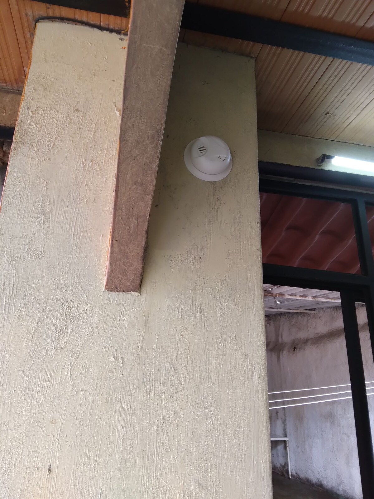 A portion of a wall is shown with a light fixture mounted near the ceiling. The surface has a textured finish in a light beige color, while the upper edge of the image reveals part of the ceiling with visible wood paneling.