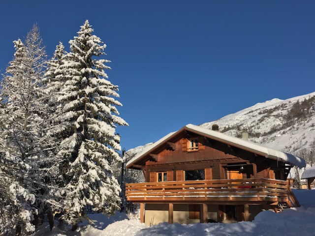A cozy chalet is nestled among snow-covered trees, with a wooden balcony that overlooks the winter landscape. The structure features a gabled roof and large windows, allowing natural light to fill the interior. A serene mountain backdrop completes the picturesque setting.