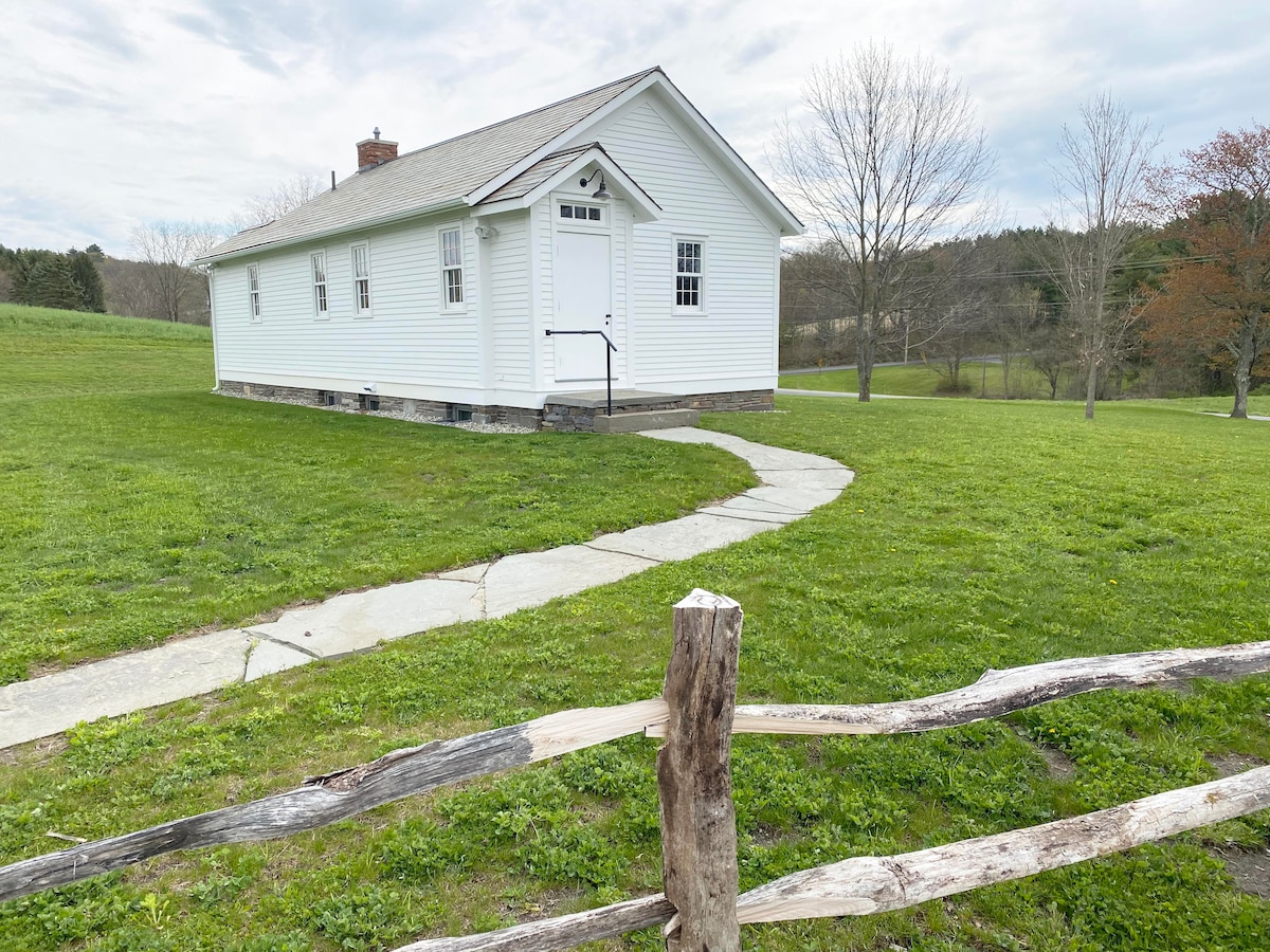 The exterior of the Buskirk School House is depicted, showcasing its classic white façade and symmetrical structure. A stone pathway leads to the front door, bordered by a wooden fence. Surrounding green lawns and trees complete the serene country setting.