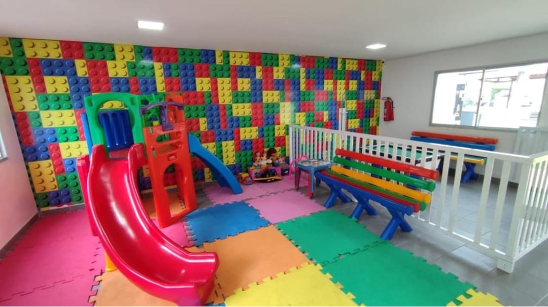 A vibrant children's play area features a red slide and colorful climbing structure, surrounded by soft play mats. Brightly colored block walls create an engaging atmosphere. Multiple benches in blue and red provide seating, while a section is cordoned off for safety.