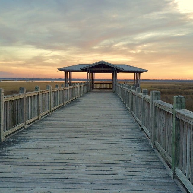 A wooden boardwalk leads to a covered gazebo, which is positioned at the end. The walkway is bordered by weathered wooden railings and stretches across a natural landscape that includes lush grasses. A soft sunset casts warm hues across the sky.