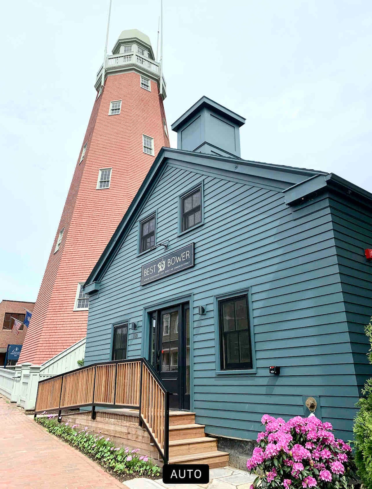 The exterior of the Best Bower guesthouse is presented with a striking blue facade accented by a wooden ramp leading to the entrance. A distinctive red brick tower is visible in the background, surrounded by vibrant pink flowers and greenery.