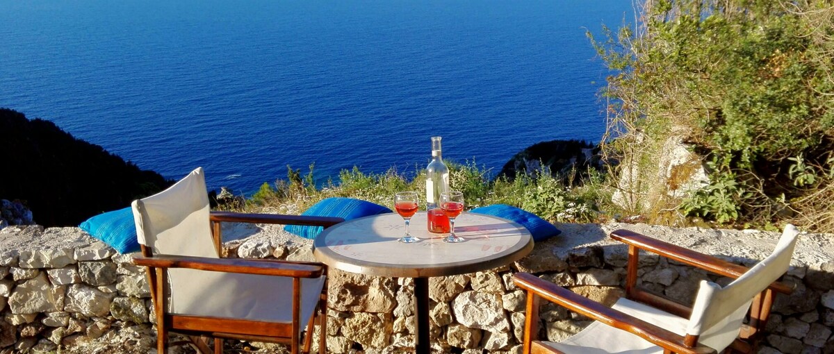 A small round table is set with glasses of red beverage and a bottle, positioned against a stone wall. Two wooden chairs with light cushions face towards a vast sea view, enhancing the outdoor setting. Green foliage frames the scene, suggesting a serene atmosphere.