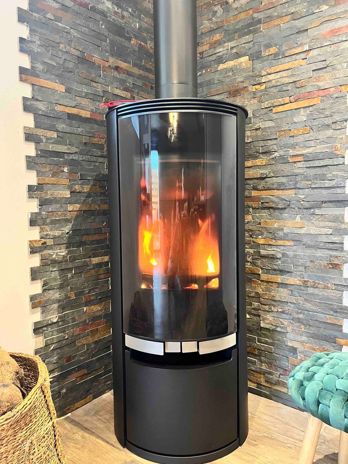 A modern black wood-burning stove is positioned against a textured stone wall, creating a warm ambiance. The glass front displays flickering flames, which add a cozy touch to the space. A woven basket is seen nearby, enhancing the rustic decor.