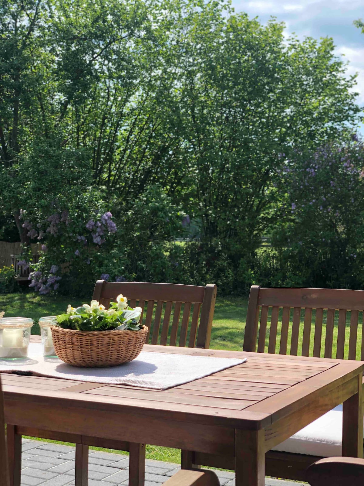 A wooden table with a light-colored tablecloth is set outdoors, accompanied by several wooden chairs. A woven basket containing potted plants and candles sits in the center. Lush greenery and flowering shrubs create a serene backdrop, enhancing the outdoor dining experience.