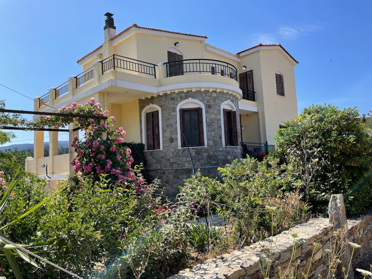 A two-story house is depicted, featuring a mix of stone and yellow exterior walls. Balconies with railings are present on multiple levels. Lush greenery and colorful flowers surround the property, enhancing the natural setting. The roof is flat, and windows are framed with wooden shutters.