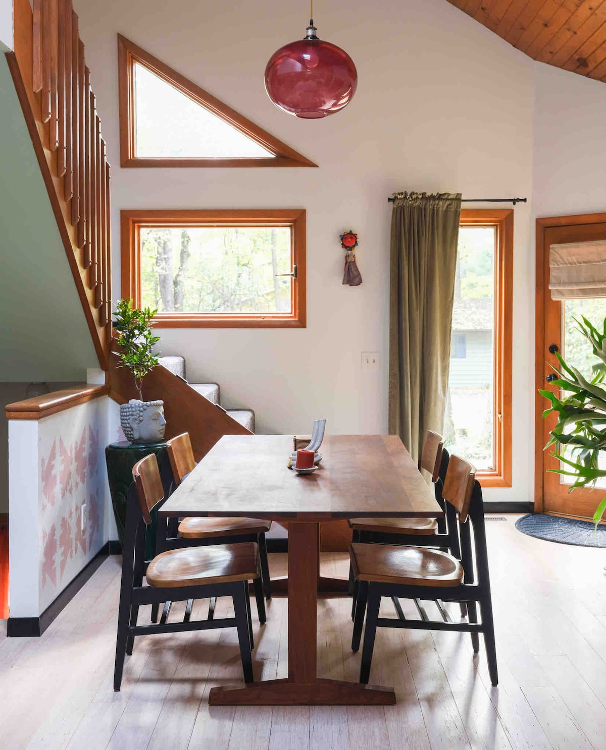 A modern dining area features a wooden table surrounded by four chairs, with natural light streaming through large windows. A decorative plant adds a touch of greenery to the space, while a pendant light hangs above, illuminating the serene atmosphere.