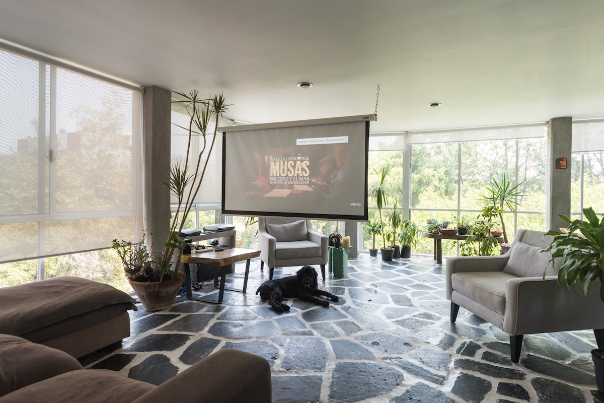 A spacious living area features a large screen for entertainment, framed by floor-to-ceiling windows that provide ample natural light. The stone floor is complemented by comfortable seating, including two armchairs. Various plants are arranged throughout the space, enhancing the connection to nature.