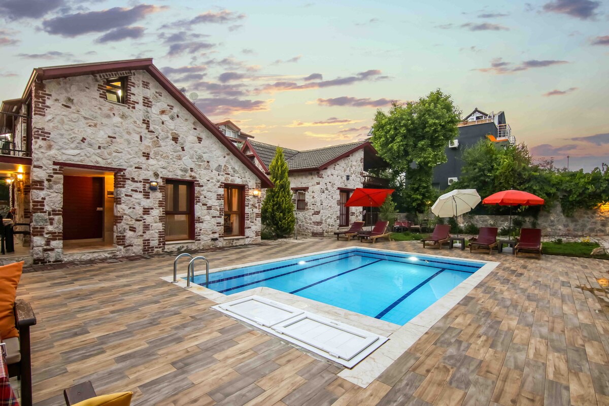 A charming villa features a rectangular swimming pool surrounded by sun loungers and shade umbrellas. The stone facade of the building complements the natural landscape, while vibrant sunset tones cast a warm light on the pool area and wooden deck.