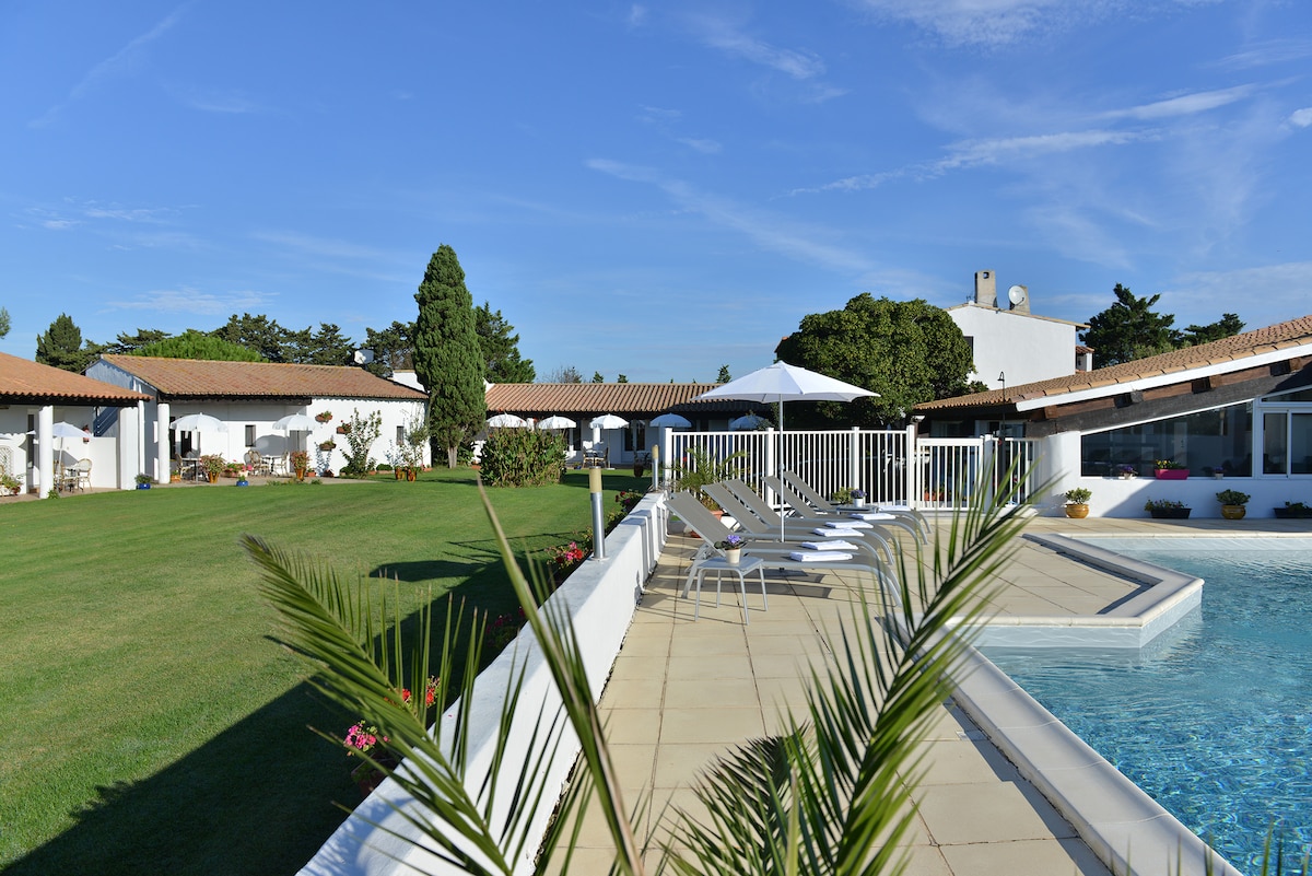 A landscaped outdoor area features a swimming pool bordered by a sun terrace with comfortable lounge chairs. The surrounding garden offers ample green space, along with mature trees and flower beds. A shaded seating area is visible, complementing the inviting ambiance.