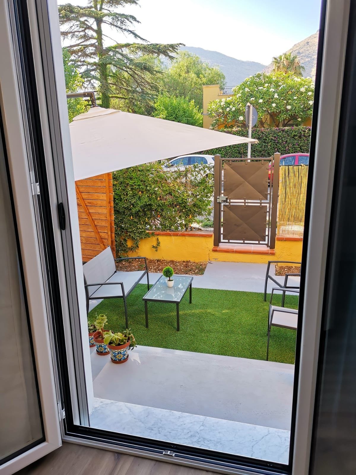 An outdoor space is visible through an open door, featuring a small grassy area with two chairs and a glass table. Potted plants add a touch of greenery, while a shaded area is created by a canopy. A gate leads to a separate section beyond.