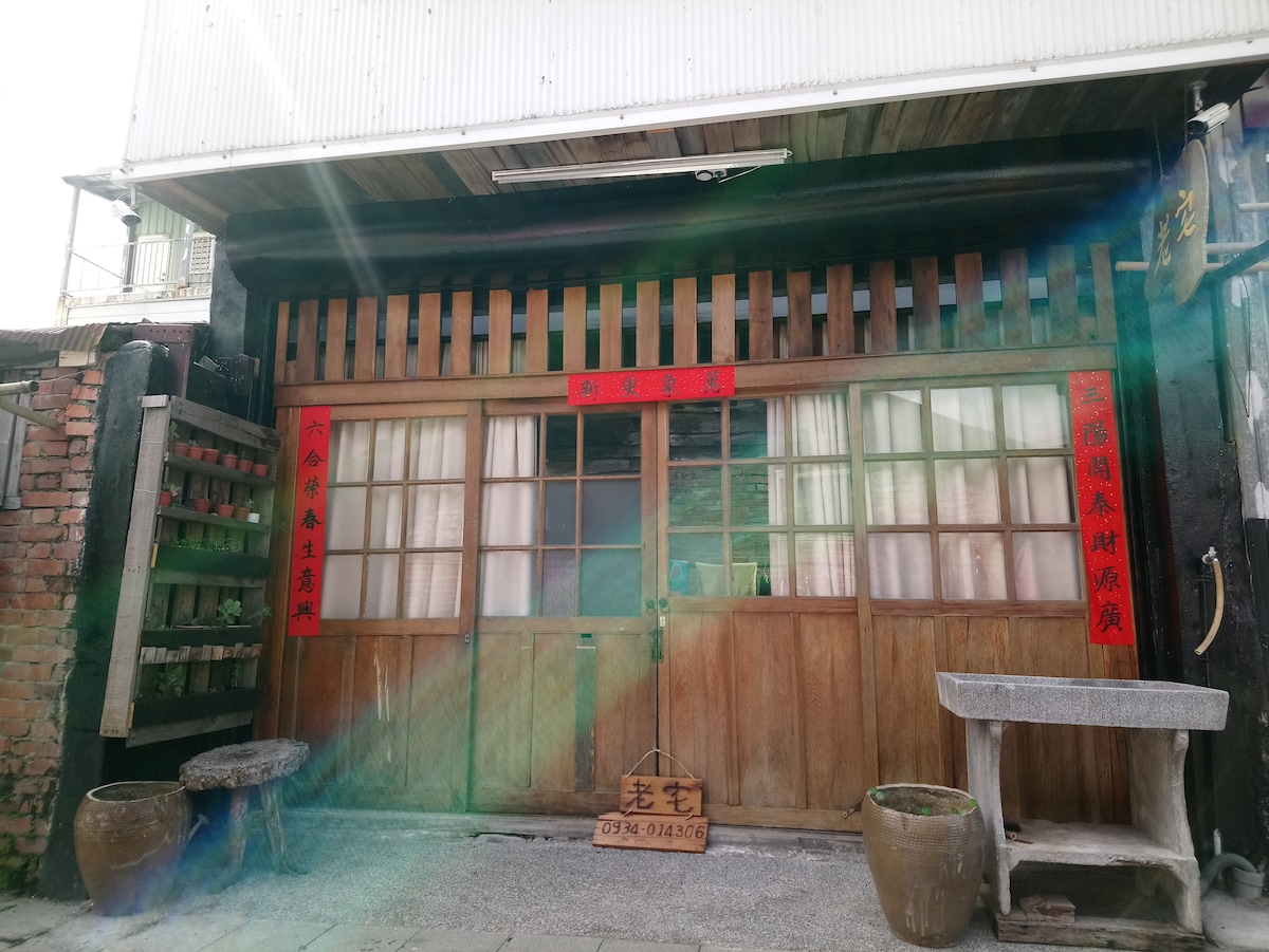 The entrance features a traditional wooden facade, showcasing intricate details. Large wooden doors are framed by vertical slats, while decorative red banners add cultural significance. Stone benches flank the entrance, enhancing the rustic charm of the building's exterior.