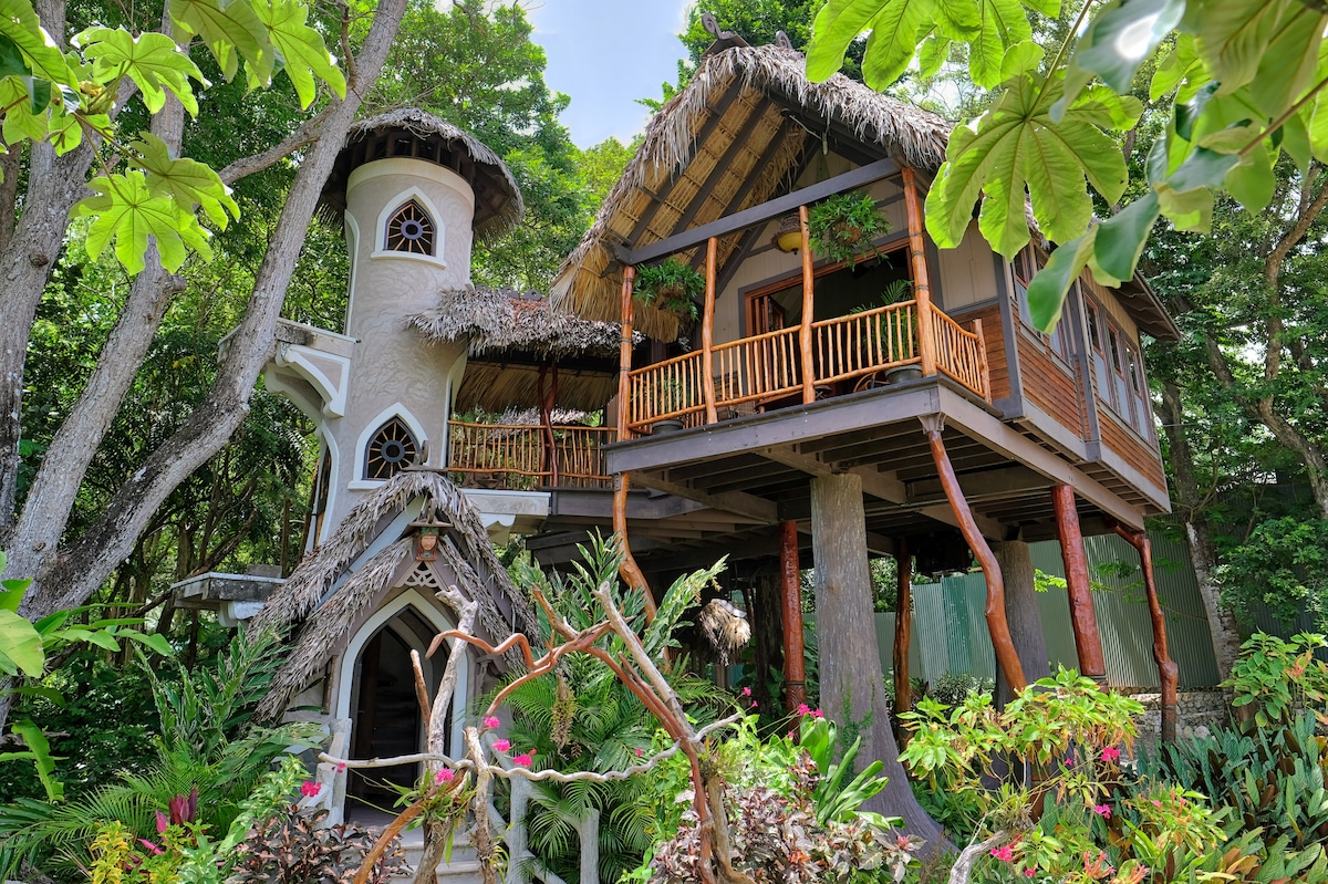 A charming tree house structure, elegantly designed with natural materials, features a thatched roof and wooden balconies surrounded by lush green foliage. The unique architecture includes a round tower and intricately crafted entryway, blending harmoniously with the vibrant tropical landscape.