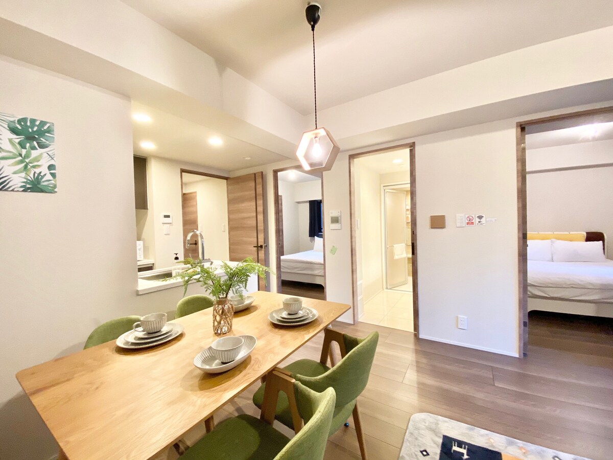 A dining area features a wooden table set for four, accompanied by green chairs. Soft lighting is provided by a geometric pendant above. Doorways lead to a well-equipped kitchen and a bedroom area, while neutral tones create a calm and welcoming environment.