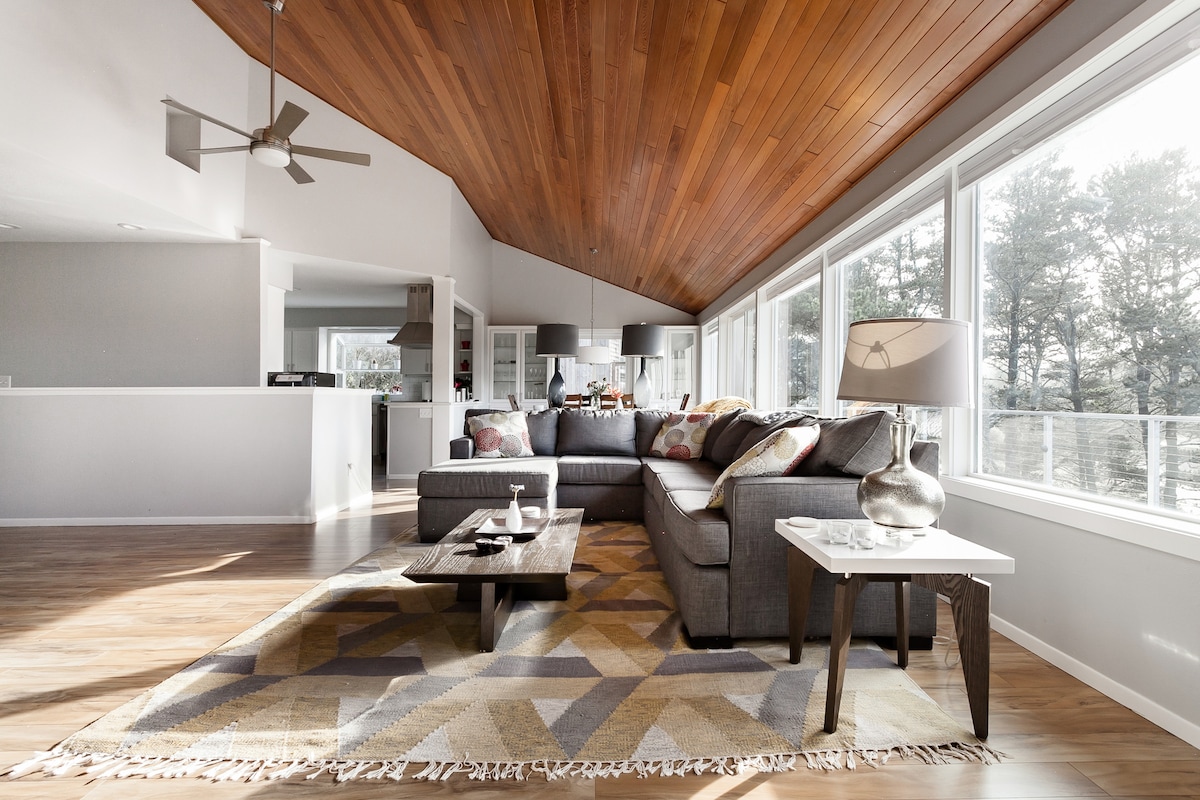 A modern living area features a spacious sectional couch facing large windows that provide a view of the surrounding woods. A wooden ceiling enhances the airy feel, while a geometric patterned rug and coffee table add warmth and texture to the space.