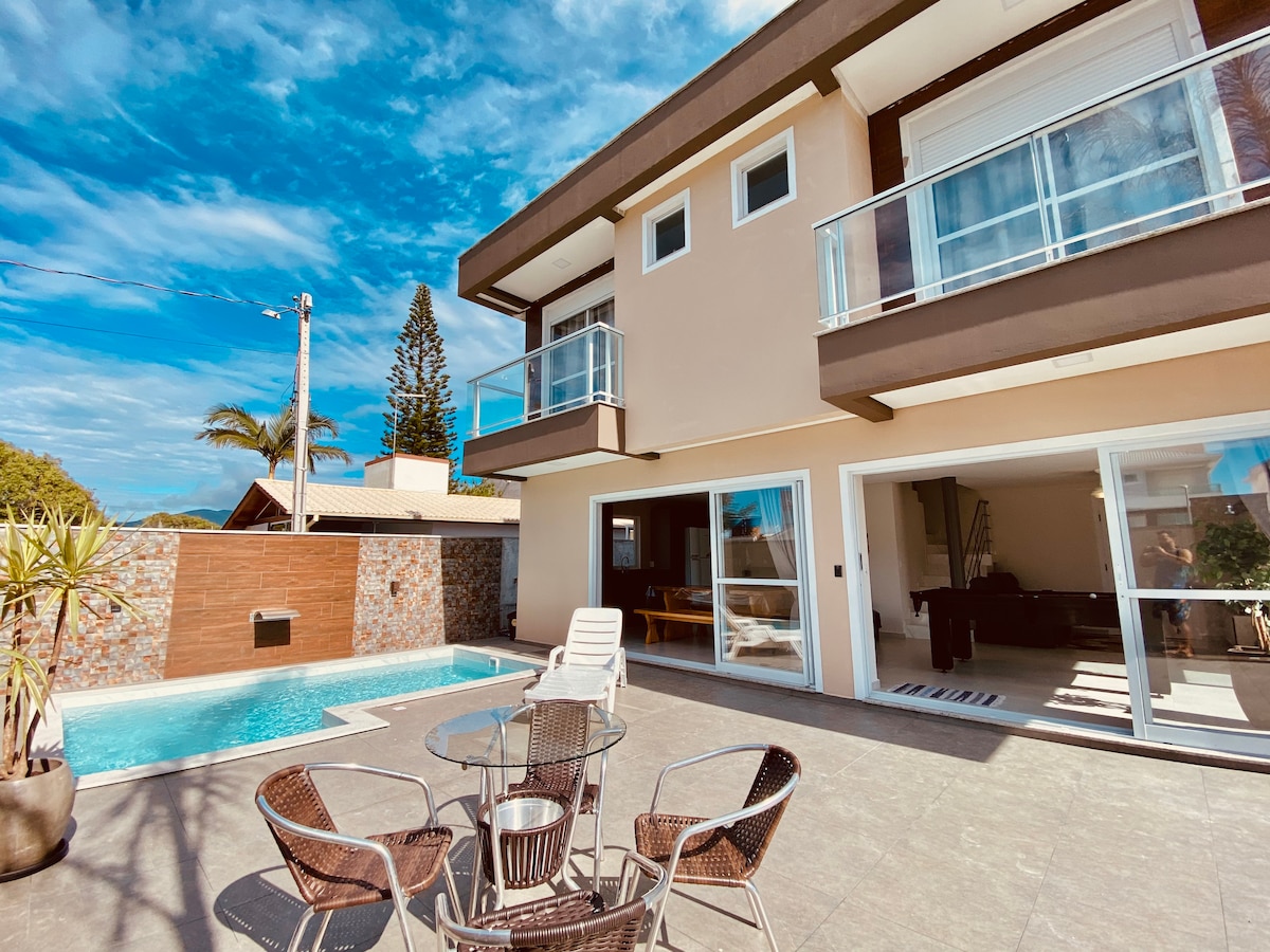 An outdoor area features a private pool alongside a spacious patio. Seated at a circular table, four wicker chairs surround the table. Large glass doors provide access into the indoor living spaces, which are visible in the background under a partially cloudy sky.
