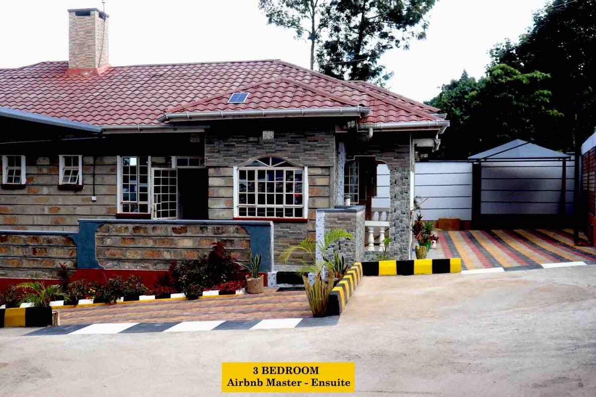 A well-designed stone house is featured, showcasing a section of the paved driveway. Colorful plants line the edge of the pathway, and a spacious parking area is visible, accommodating multiple vehicles. The roof is covered with red tiles, and windows are adorned with decorative grills.