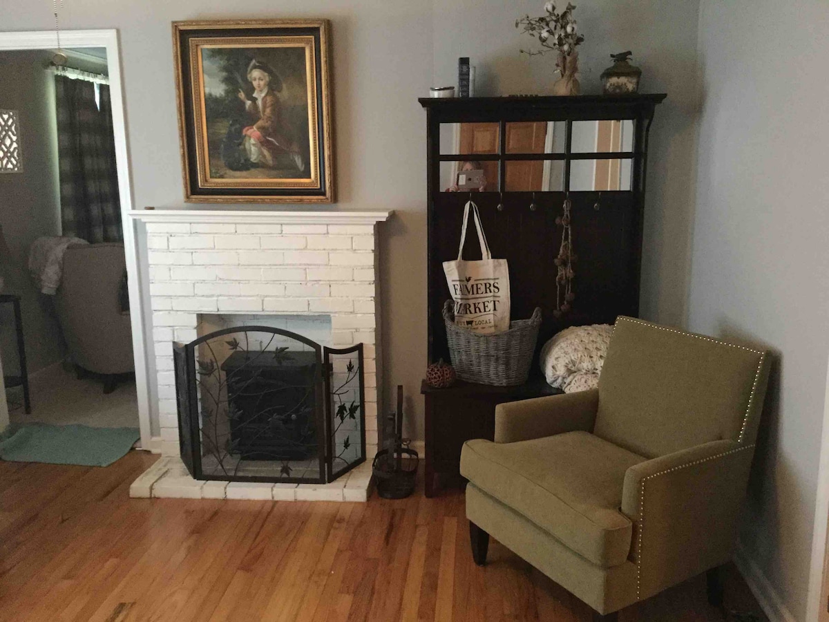 A cozy living area features a white brick fireplace with a decorative screen, complemented by an elegant painting above. A green accent chair sits nearby, alongside a dark wooden cabinet with hooks and a decorative bag. Warm wood flooring adds to the inviting atmosphere.