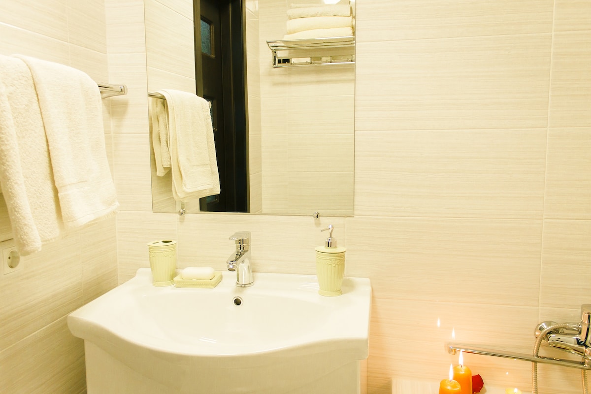 A bright, modern bathroom is displayed, featuring a curved white sink and a large mirror. Soft white towels hang neatly on a rack, and small containers for toiletries are placed on the counter. A lit candle adds a gentle glow to the space.