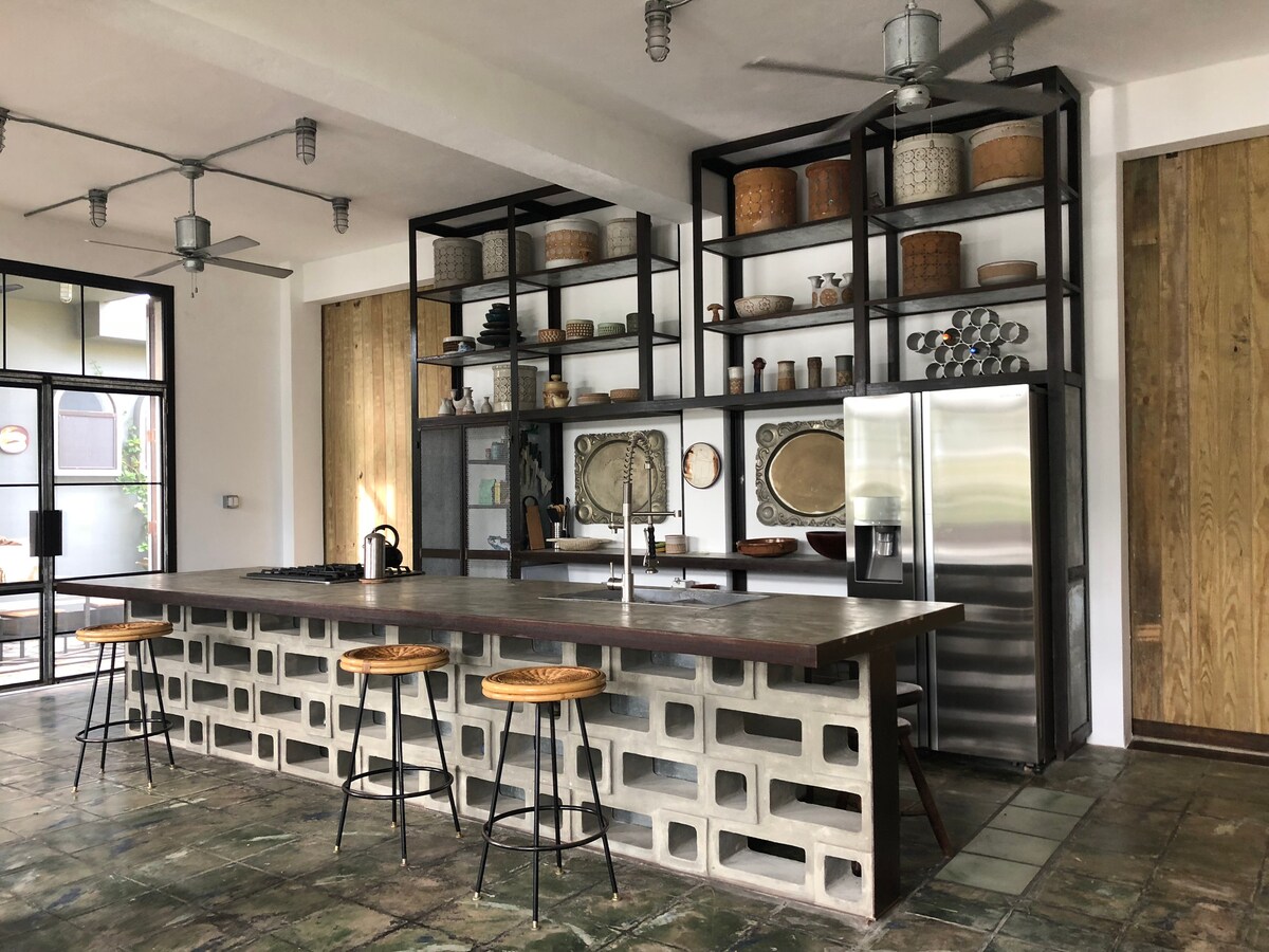 A spacious kitchen features an expansive island with bar stools, surrounded by custom shelving displaying decorative items. Stainless steel appliances are integrated into the design, while industrial fans provide airflow, contributing to the open atmosphere. Large windows offer natural light and a view of the outdoor area.