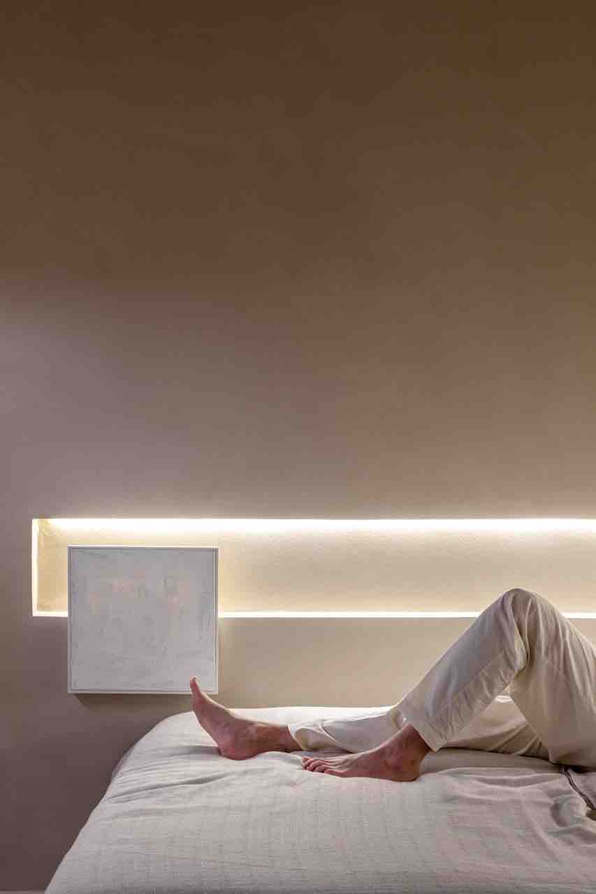 A spacious bed is positioned against a softly lit wall, creating a serene atmosphere. A subtle LED light strip runs along the headboard, enhancing the contemporary design. Light fabric covers the bed, inviting relaxation. A pair of feet can be seen, emphasizing comfort.