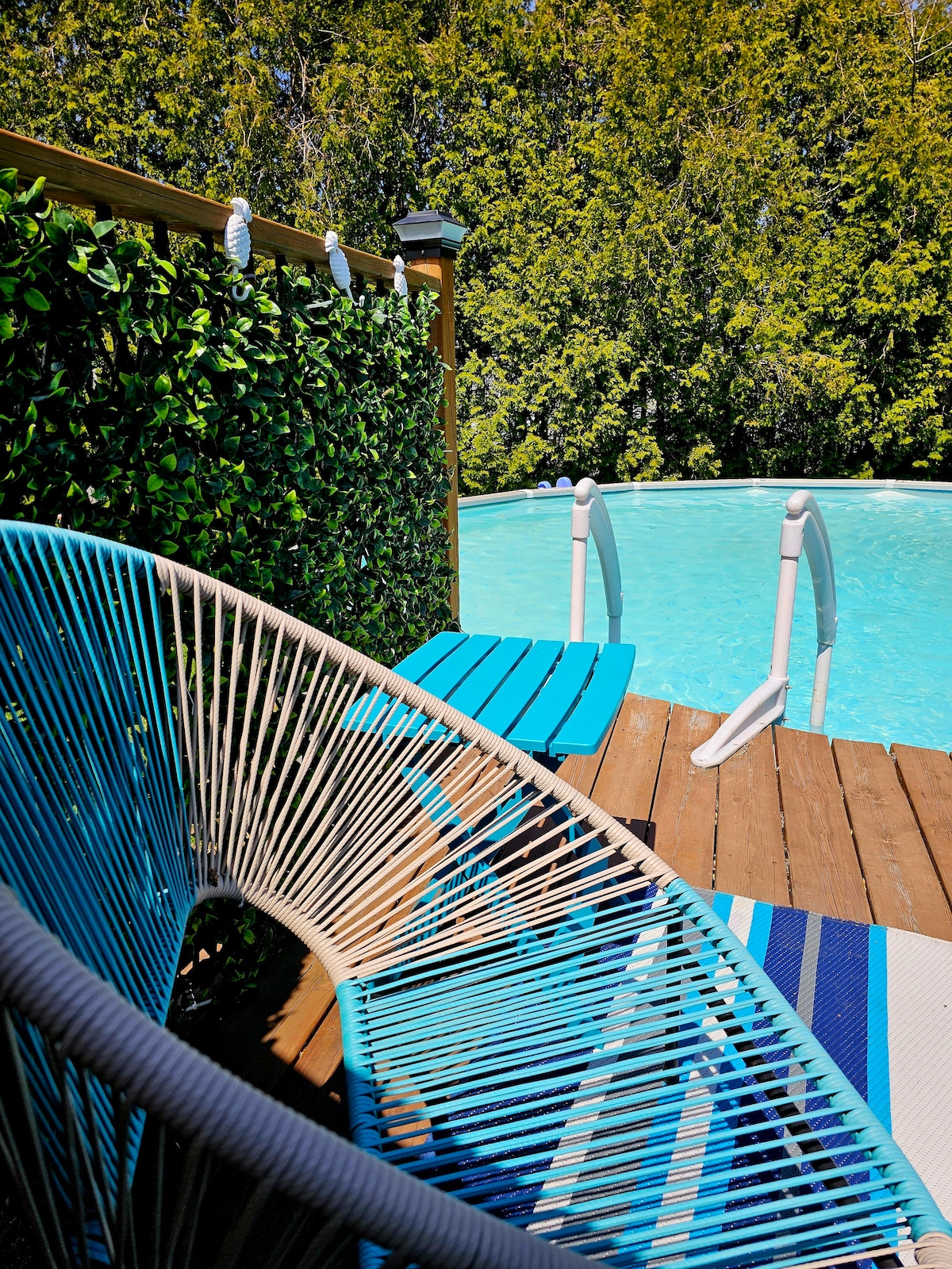 A refreshing pool is surrounded by a wooden deck, featuring a bright blue lounge chair and a striped area rug. Lush greenery provides privacy, enhancing the serene outdoor space available for relaxation.