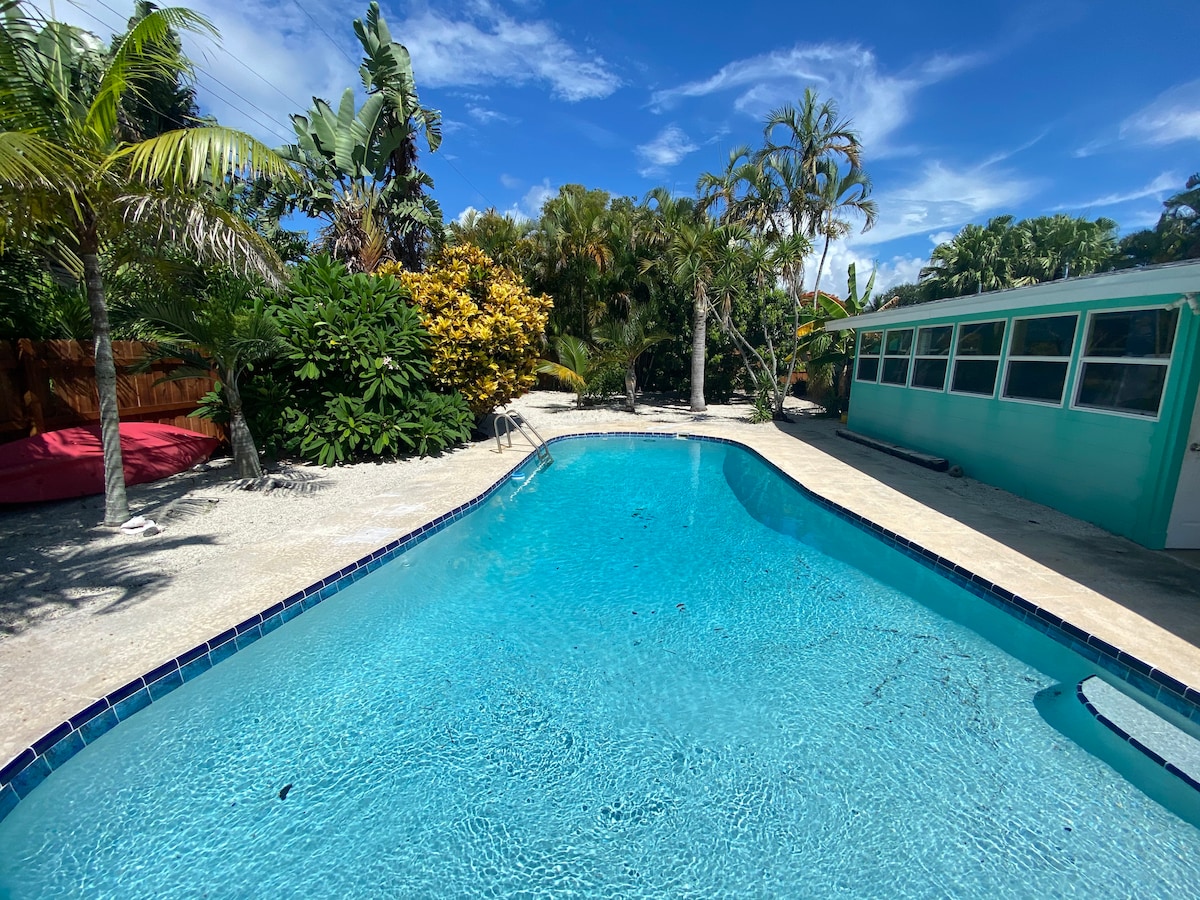 A large pool is surrounded by tropical foliage and fruit trees, creating a refreshing oasis. The bright blue water reflects the clear sky, while the pool area features a spacious deck suitable for relaxation. The vibrant greenery provides a natural backdrop.