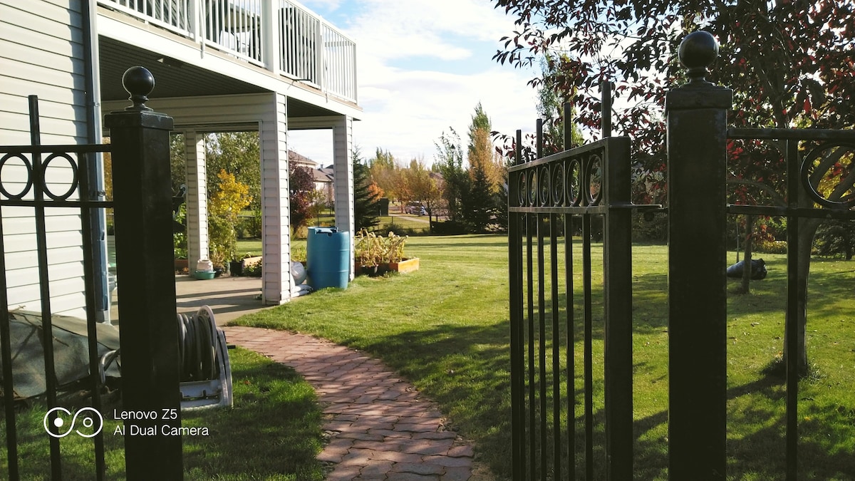 A gated entrance leads to a well-maintained path winding through a lush green yard. Beyond the gate, an expansive lawn is visible, bordered by trees and shrubs, creating a serene outdoor space with a peaceful atmosphere.