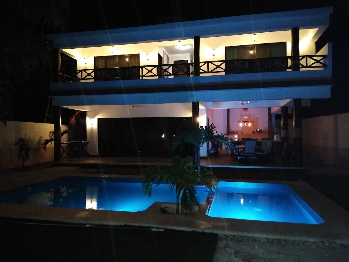 The two-story house is illuminated at night, displaying a modern architectural design. A pool reflects soft blue light, surrounded by lush greenery. Sliding glass doors reveal a cozy indoor space, while the upper balcony features dark wooden railings and outdoor seating.