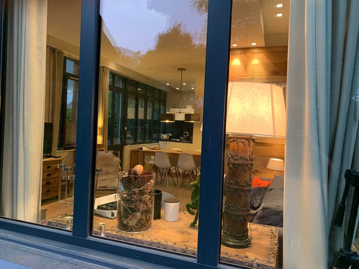 A view through expansive glass windows reveals a cozy living space, featuring a large lamp and a wooden table surrounded by stylish chairs. Natural light filters through the curtains, highlighting a relaxed ambiance complemented by decorative elements, including woven baskets and a plush sofa.