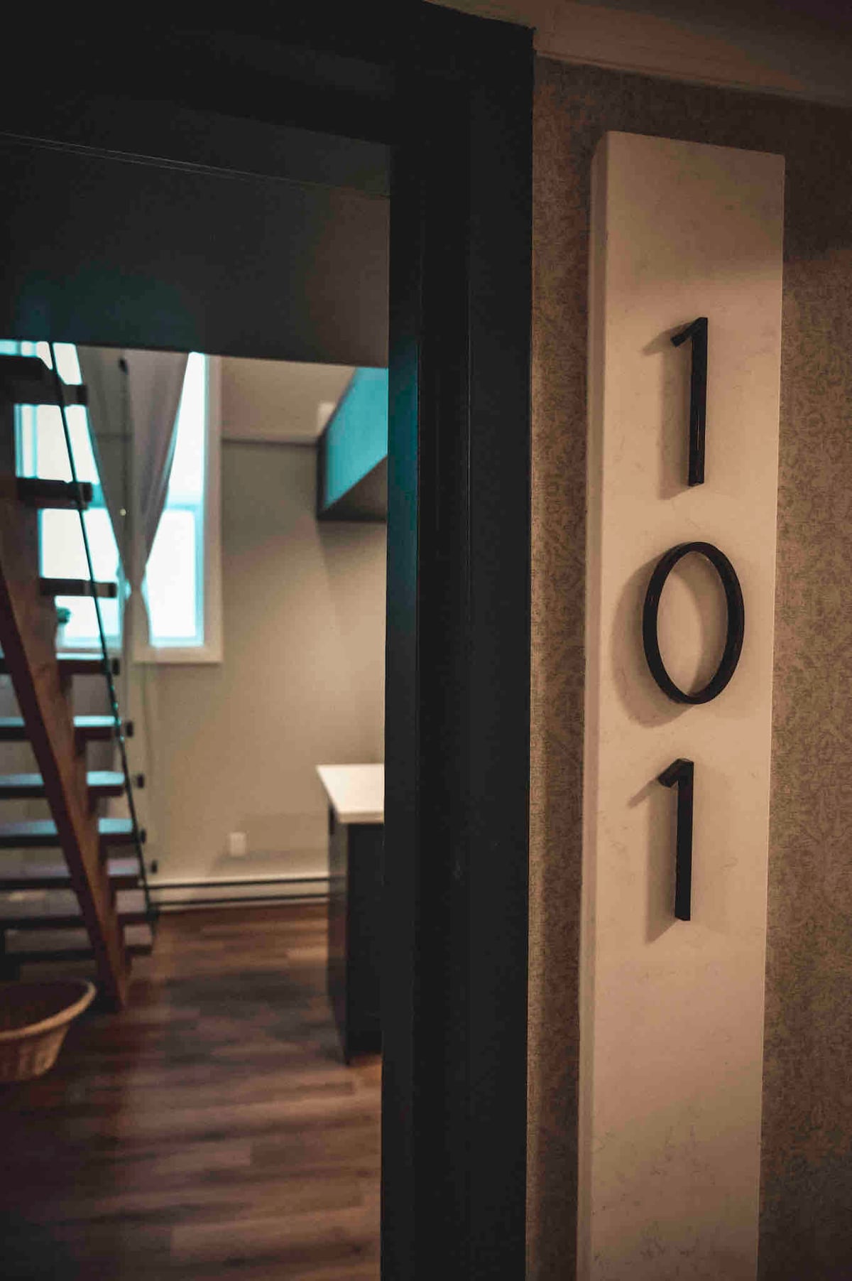 A doorway is framed by a dark entrance, featuring a clear view of an apartment number '101'. A wooden staircase is visible in the background, leading to an upper level, while light enters through a window, illuminating the warm wood flooring.
