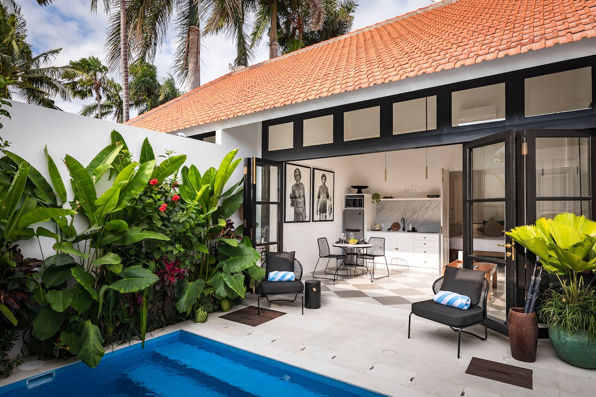 A private outdoor area showcases a crystal-clear pool surrounded by tropical plants. A seating area is arranged with several lounge chairs and a small table, inviting relaxation. The villa features large glass doors that open to a spacious living area adorned with modern decor.