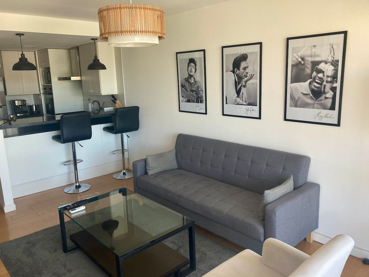 A modern living area features a gray sofa and a glass coffee table, complemented by two bar stools at a nearby countertop. Black and white framed artwork adorns the walls, adding character to the space. Natural light illuminates the room through a large window.