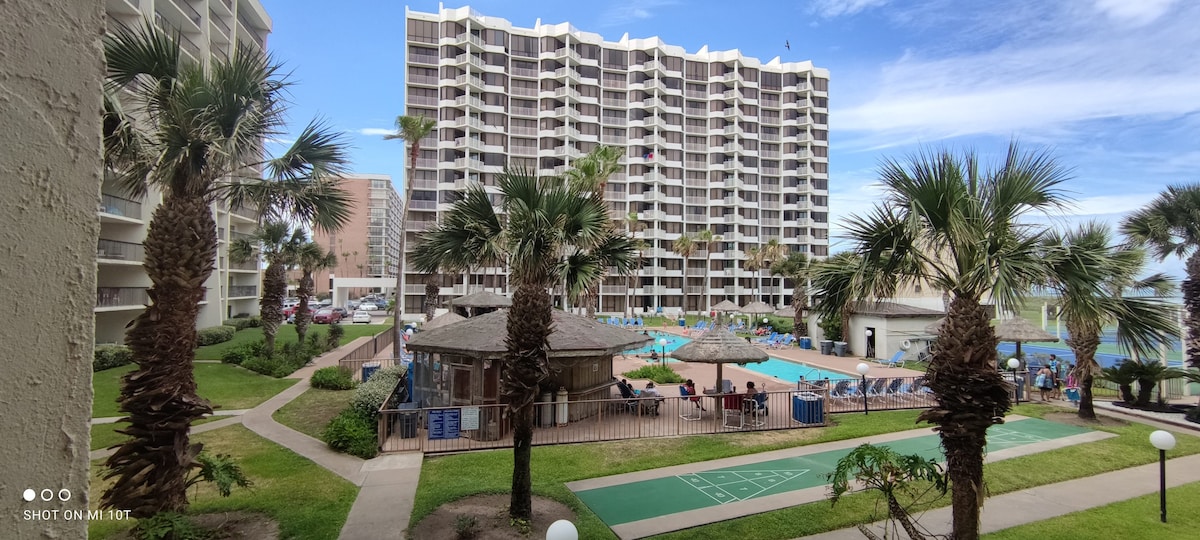 The view from the balcony captures a landscaped area with palm trees and a pool surrounded by loungers. The distant building showcases multiple levels, while shaded seating areas near the pool enhance the outdoor experience. Clear skies are seen above, contributing to the inviting ambiance.