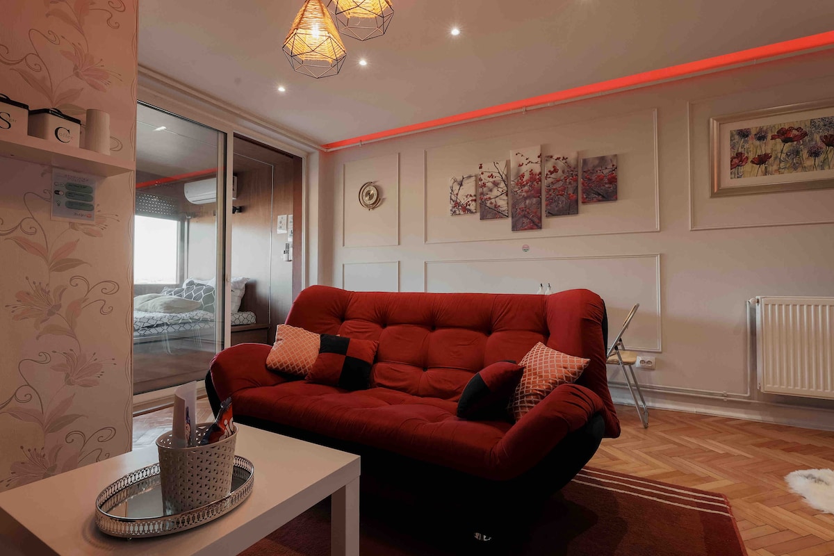 A cozy living area features a red futon with various decorative pillows and a small white coffee table. Soft lighting is provided by pendant lamps, and artwork adorns the wall. A sliding glass door leads to a bedroom area in the background, enhancing the open feel.