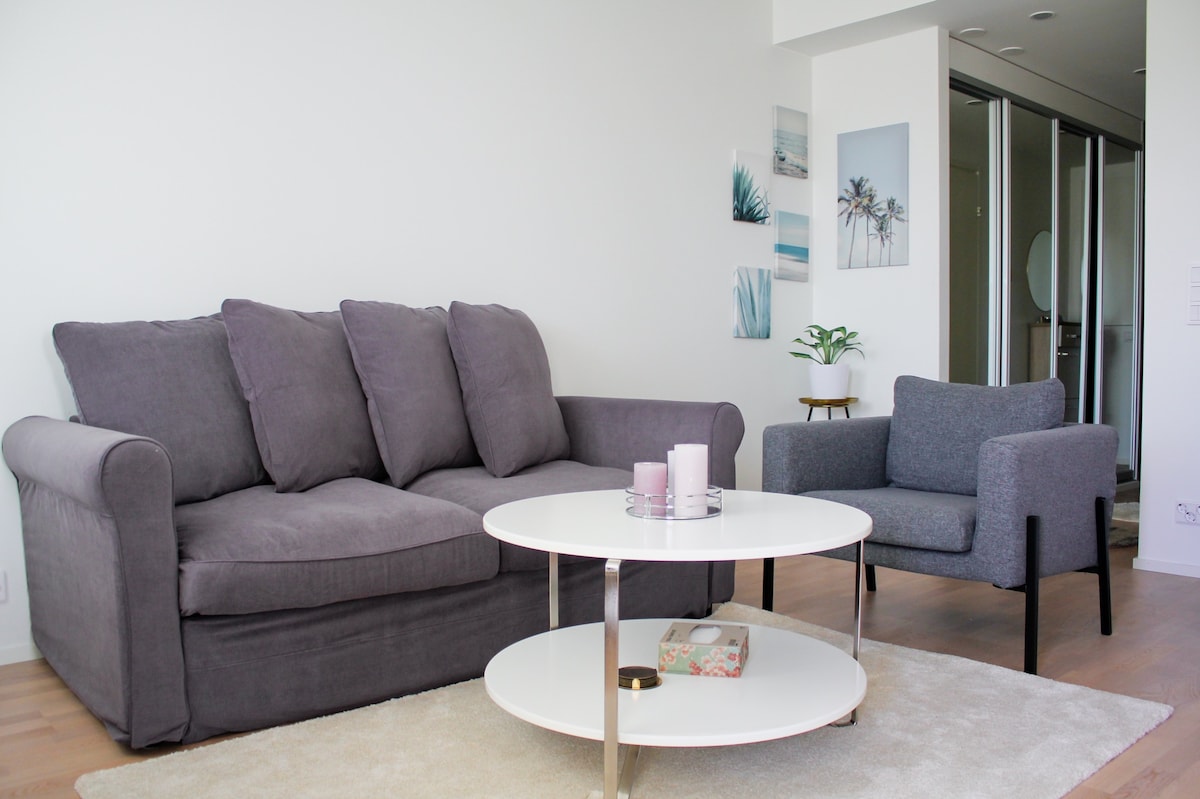 A light-filled living area features a gray sofa and a matching armchair, complemented by a round coffee table. A neutral rug lies beneath, and artwork depicting serene beach scenes adorns the walls, enhancing the room's calm atmosphere.
